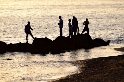 A group of people standing on rocks at the beach, silhouetted against the sea at sunset.